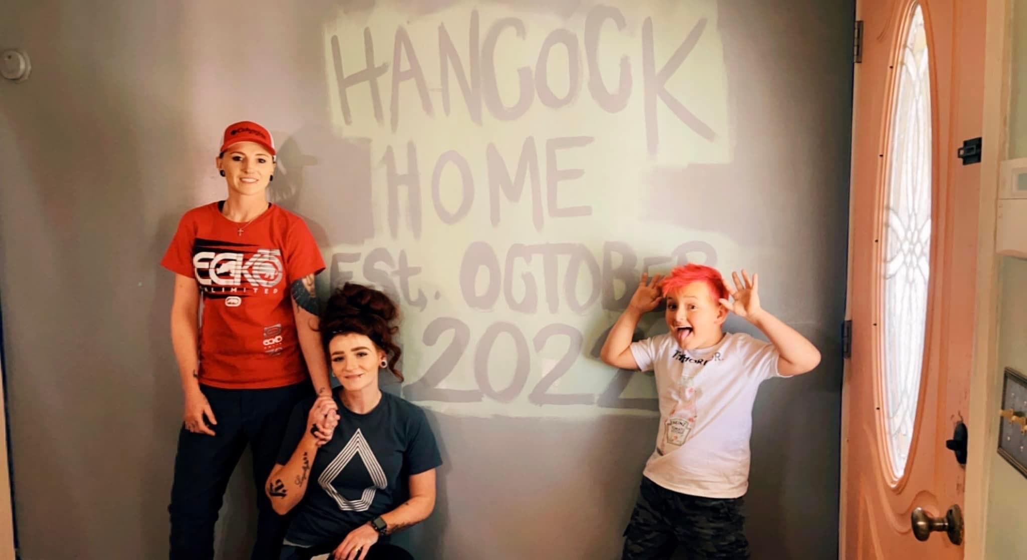 Two women and a boy with pink hair pose by a 'Hancock Home' wall.