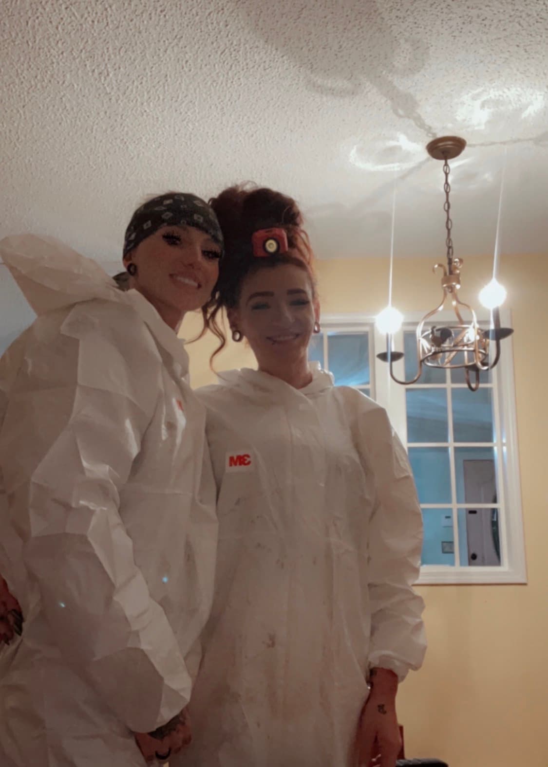 Two smiling women in white protective suits, one wearing a headlamp and one a bandana.