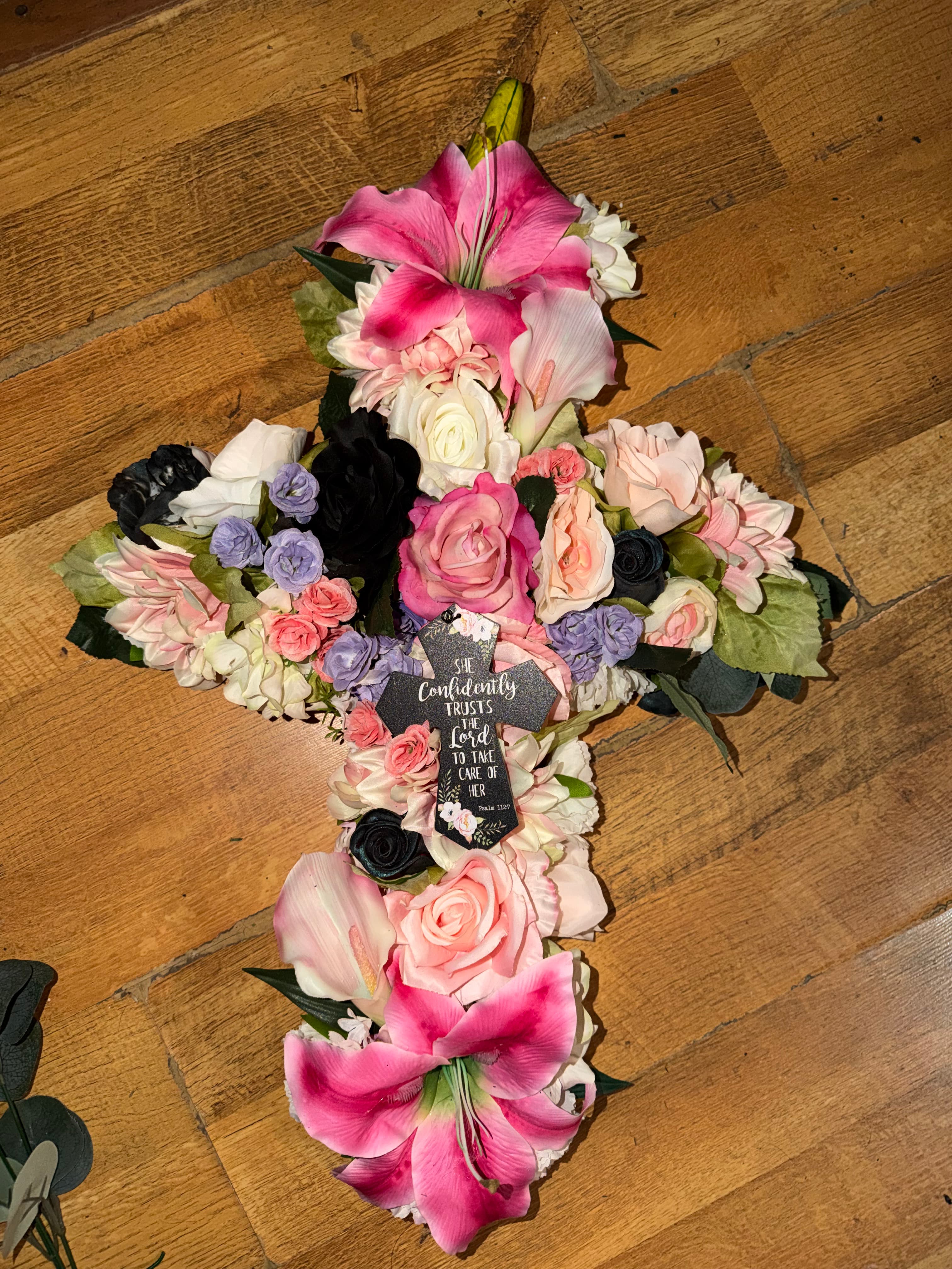 Cross-shaped floral arrangement with pink, white, and black flowers and a central religious plaque.