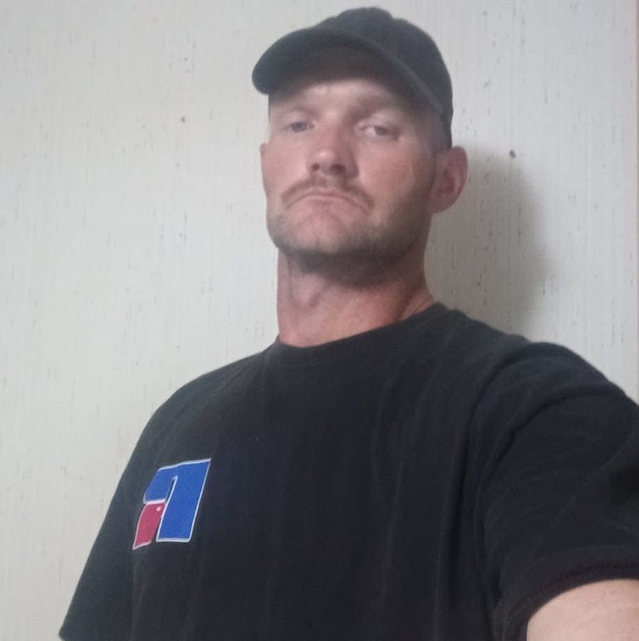 Man with a mustache and goatee wearing a black baseball cap and black t-shirt.