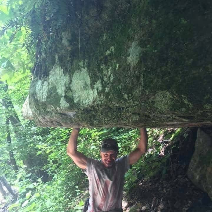 Smiling man in a lush forest pretending to hold up a massive mossy rock overhang.