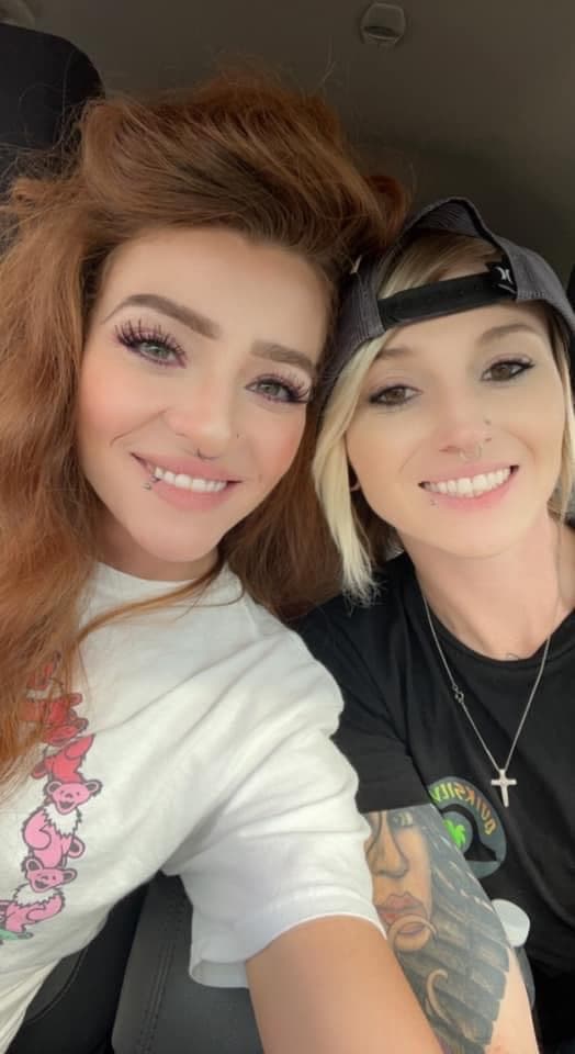 Two women with facial piercings and a tattoo smile for a selfie inside a car.