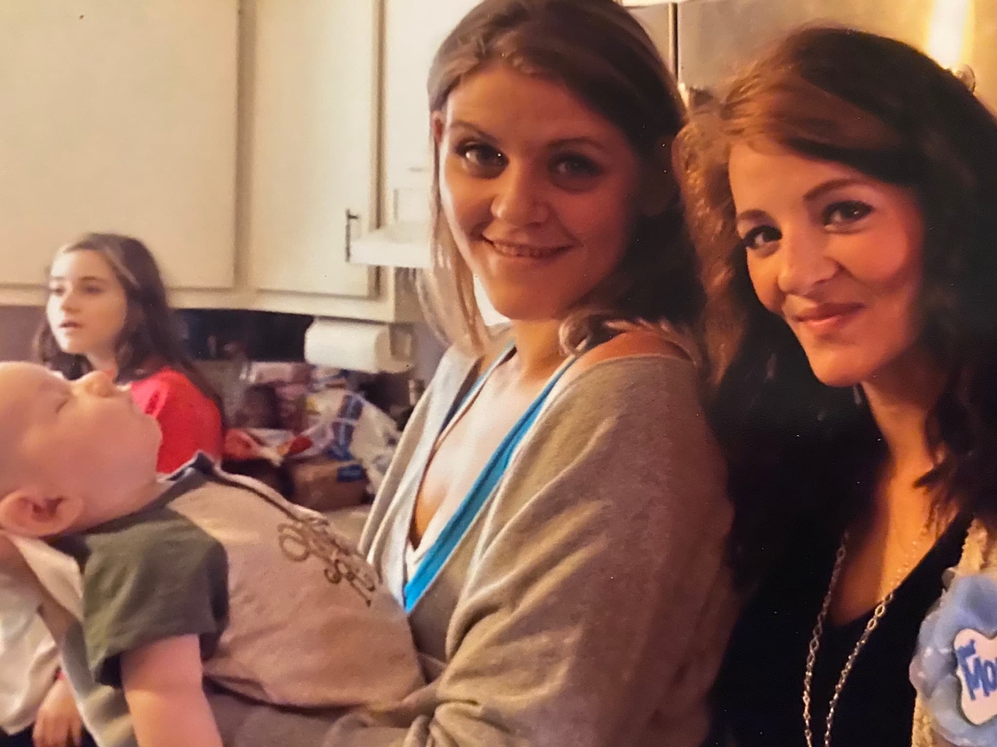 Two smiling young women pose in a kitchen while one holds a sleeping baby.
