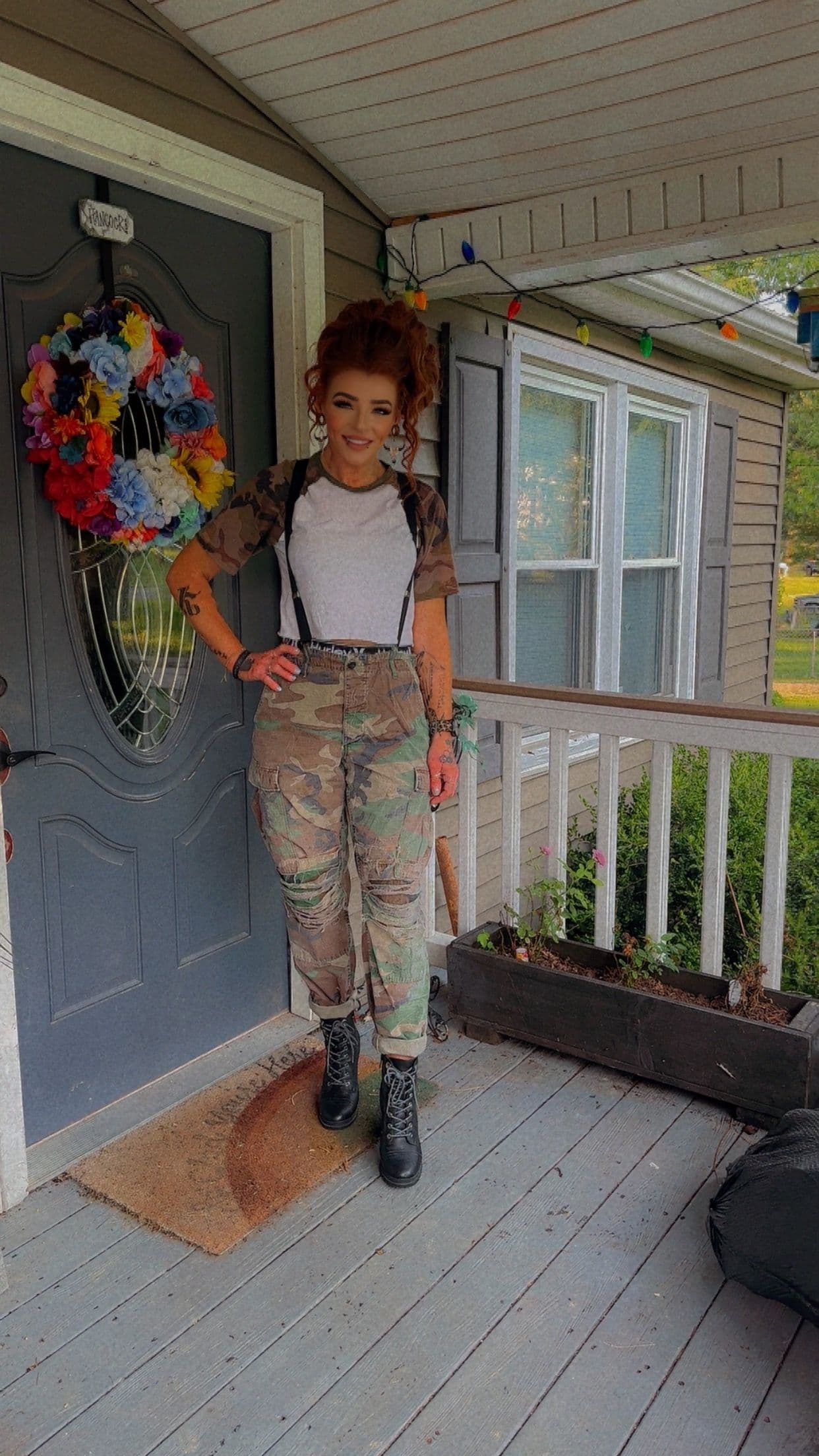 Red-haired woman in camouflage pants and suspenders stands before a blue door with a wreath.